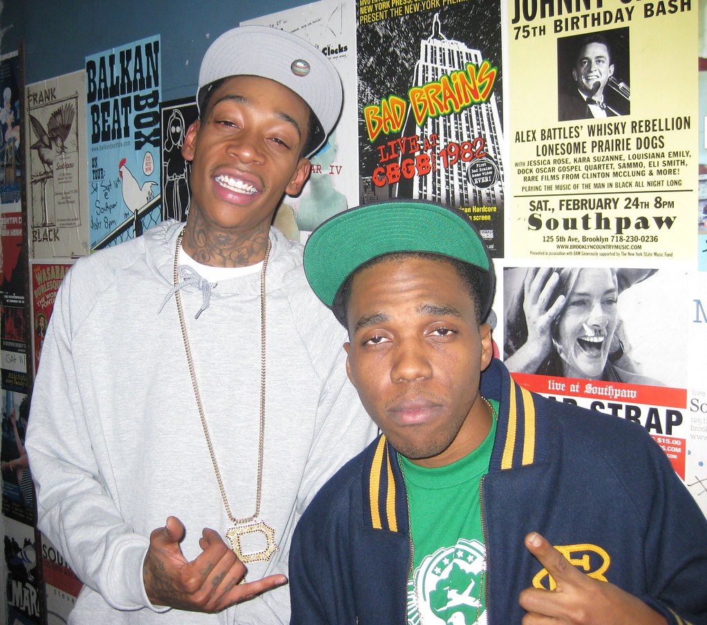 Wiz Khalifa smiles beside Curren$y in a varsity jacket backstage at Southpaw, Brooklyn, in front of concert posters on 20 October 2009.